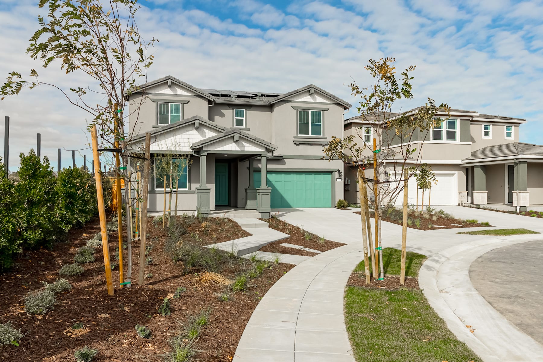 A well-landscaped residential neighborhood with a two-story house featuring a green garage door, surrounded by trees and a paved walkway leading to the entrance.