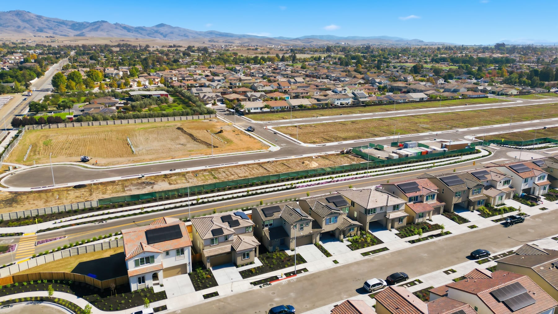 A sprawling residential neighborhood nestled among rolling hills, with a railway line running through the foreground and a vast expanse of open fields in the background, all under a clear blue sky.