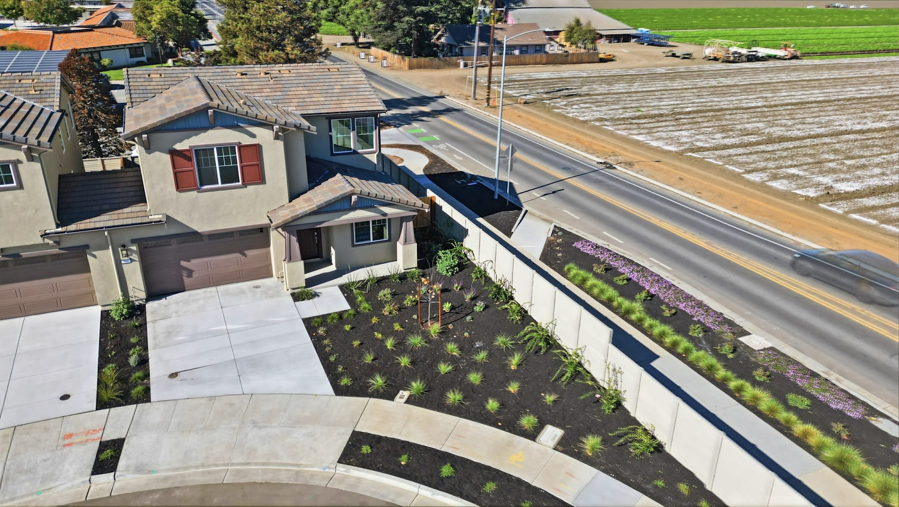 A residential neighborhood with a winding path, landscaped gardens, and a view of agricultural fields in the background.