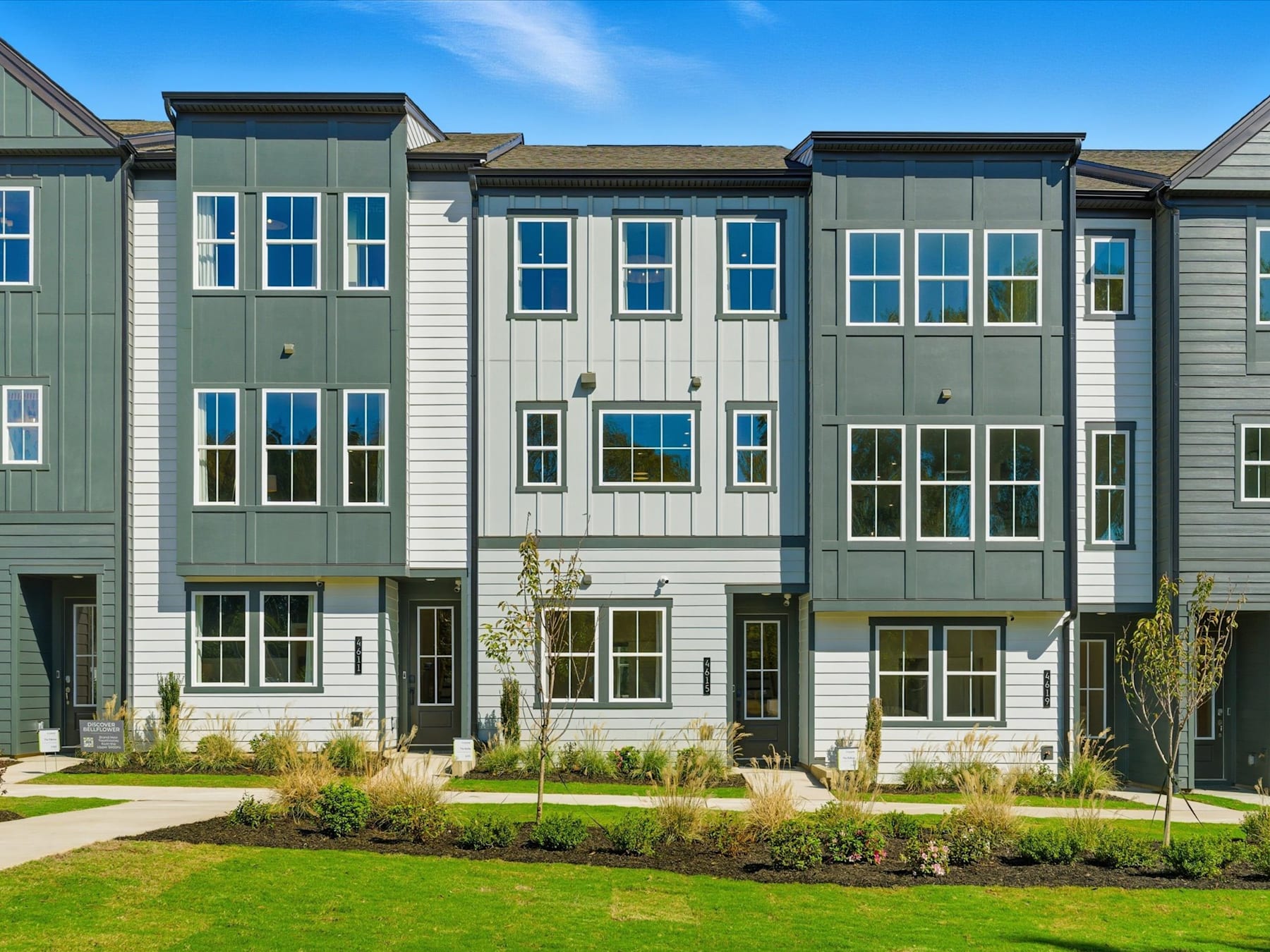 A row of modern, multi-story townhouses with green siding, white trim, and large windows, set against a backdrop of a clear blue sky and surrounded by a well-manicured lawn.