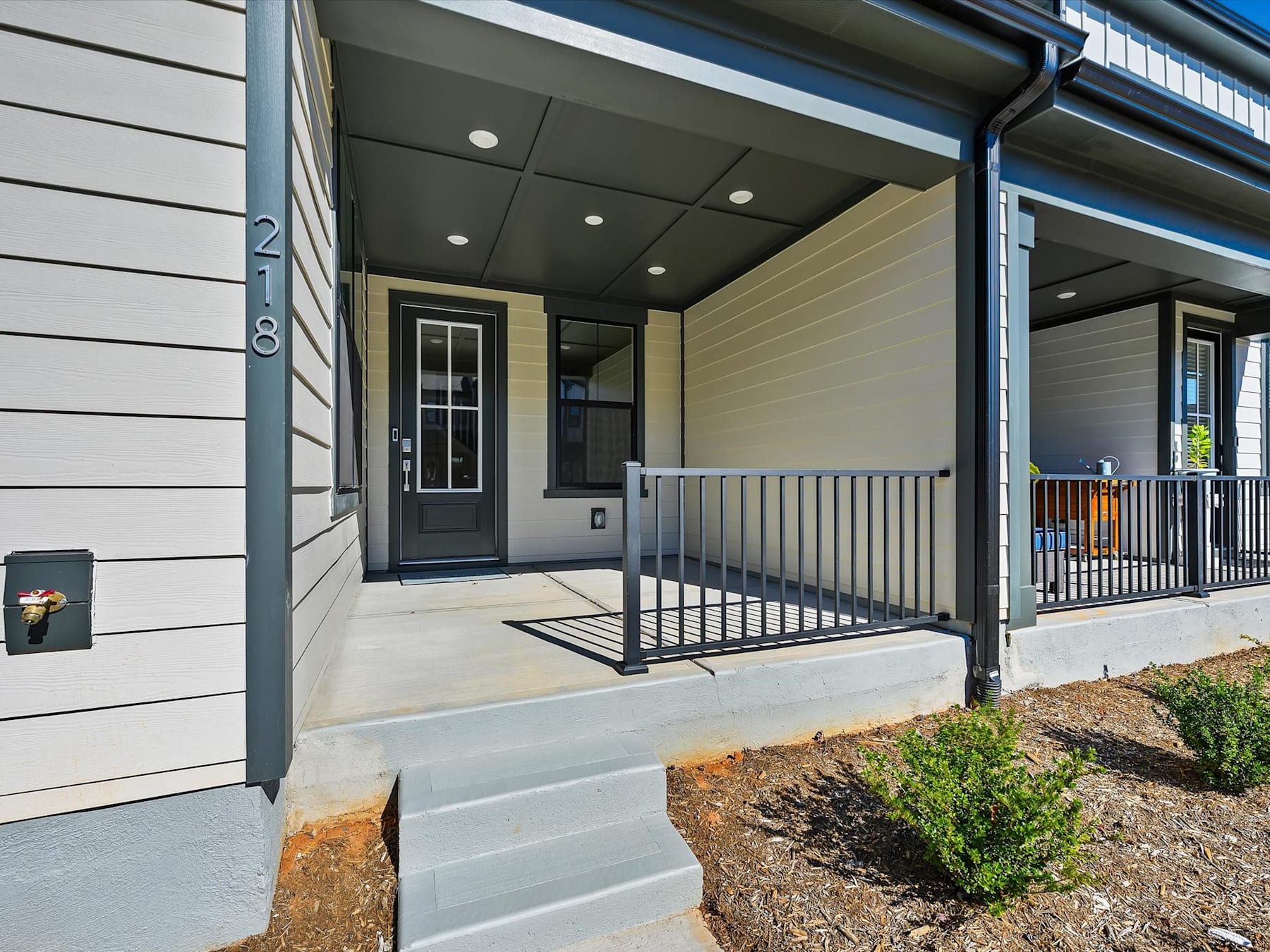 The image shows the exterior of a modern, two-story residential building with a covered porch, wooden siding, and a metal railing leading up to the front door.
