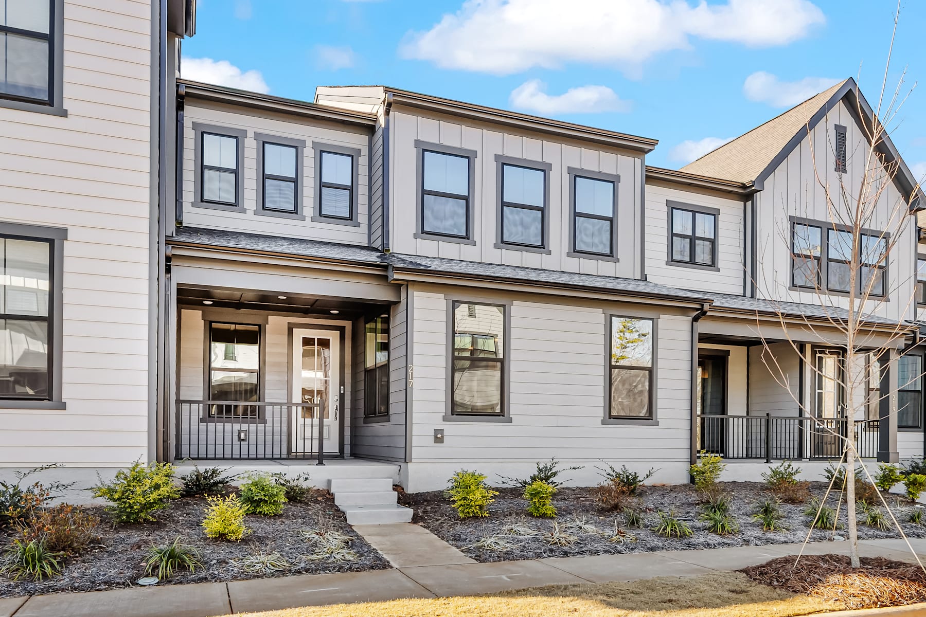 A multi-story residential building with a gray exterior, featuring large windows, a covered entryway, and a landscaped front yard with plants and gravel.