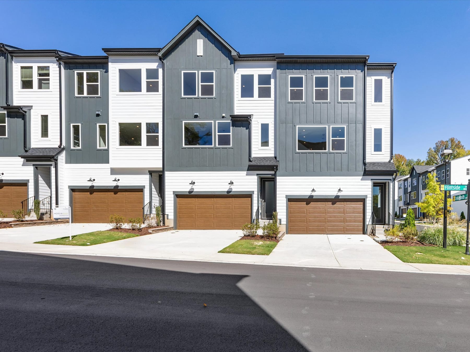A row of modern, multi-story townhouses with gray and white siding, featuring garages and landscaped yards in the foreground, set against a clear blue sky in the background.