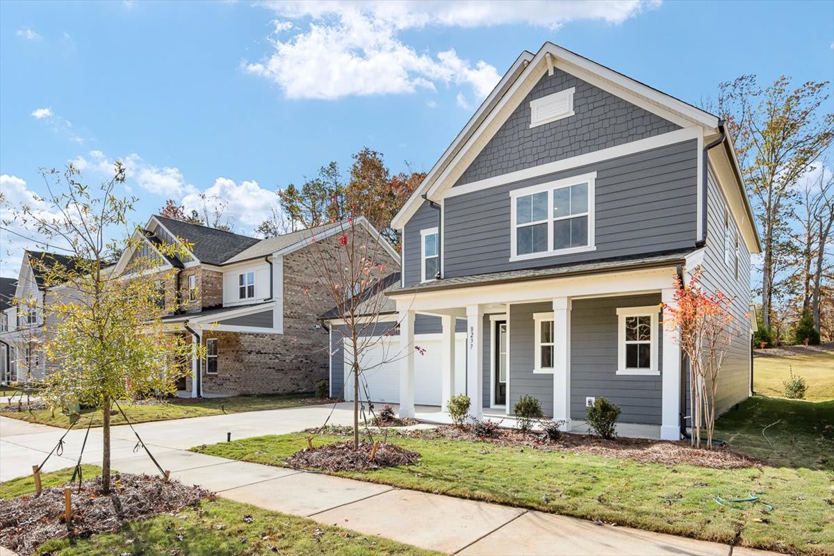 A well-maintained, two-story residential home with a gray exterior, a garage, and a neatly landscaped yard surrounded by trees in a suburban neighborhood.
