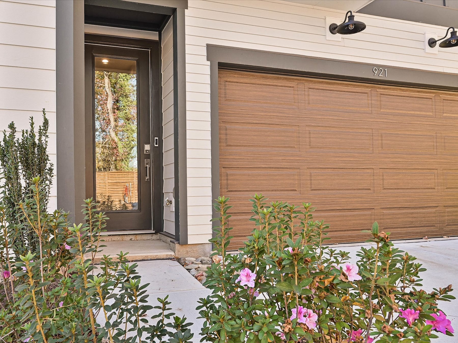 A wooden front door with a glass panel, surrounded by a lush garden with various flowering plants and shrubs, stands in the foreground of the image.