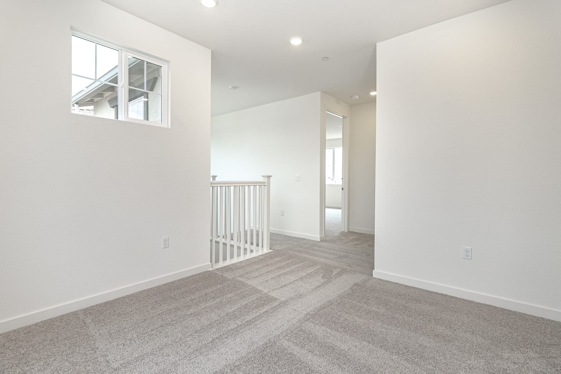 A bright, open hallway with white walls, a carpeted floor, and a window providing natural light, leading to a doorway in the background.