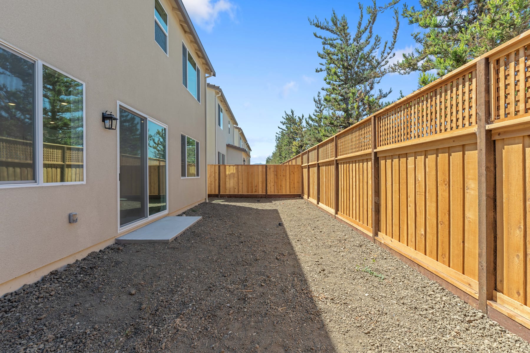 A wooden fence runs along the side of a residential building, with a gravel path leading through the backyard and surrounded by pine trees.