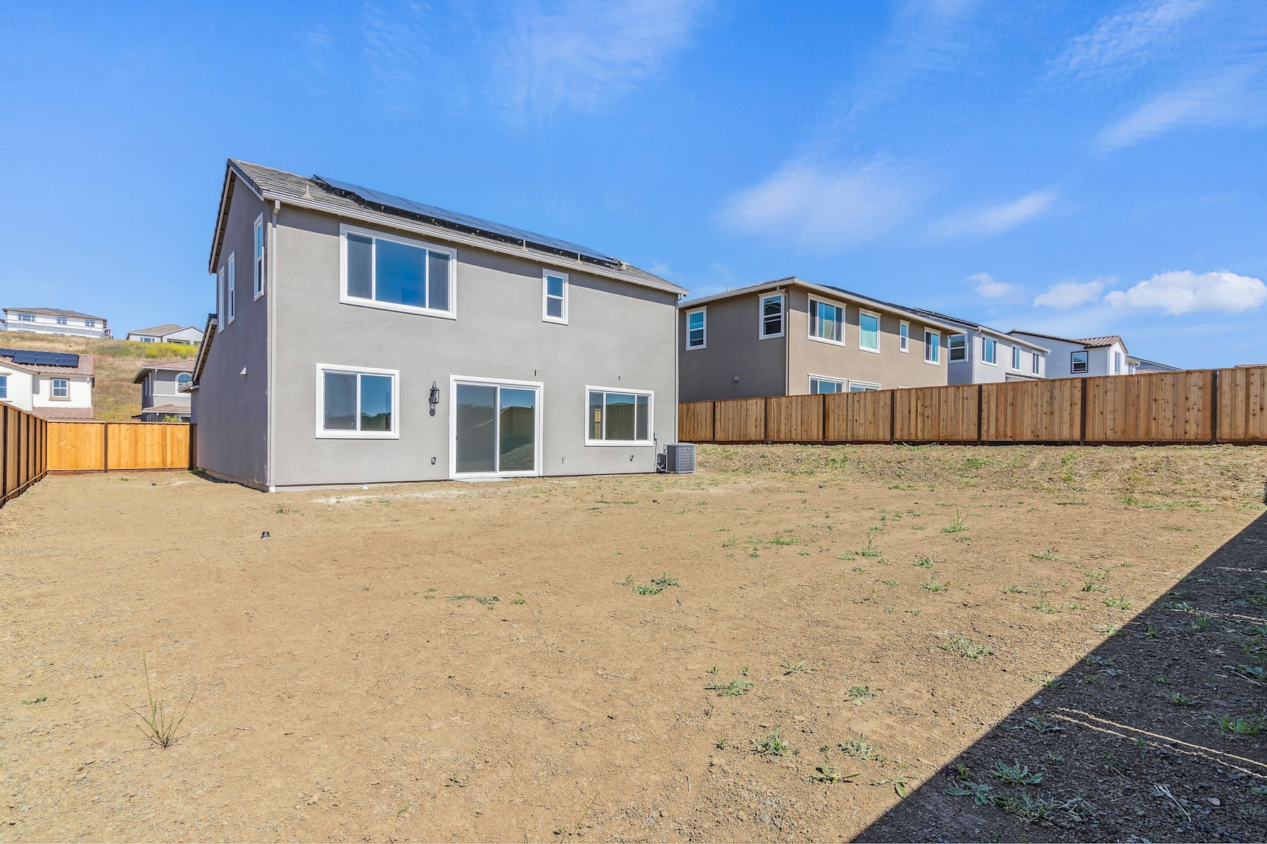 A row of modern, gray-colored townhouses with wooden fences in the foreground, set against a clear blue sky with scattered clouds.