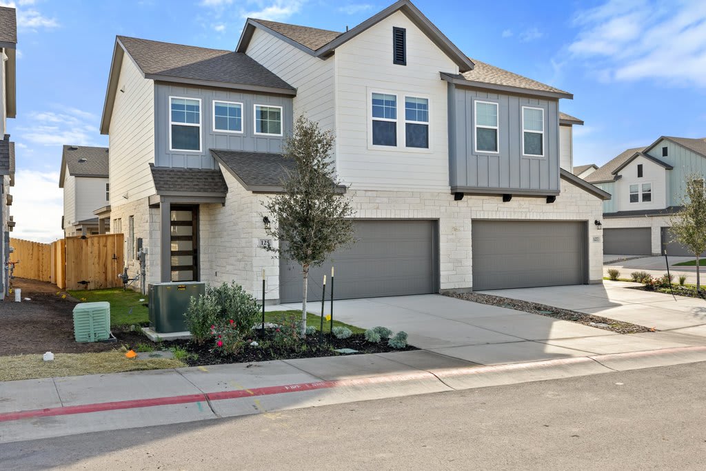 A two-story residential house with a garage, surrounded by landscaping and a paved driveway, set against a clear blue sky with some clouds.