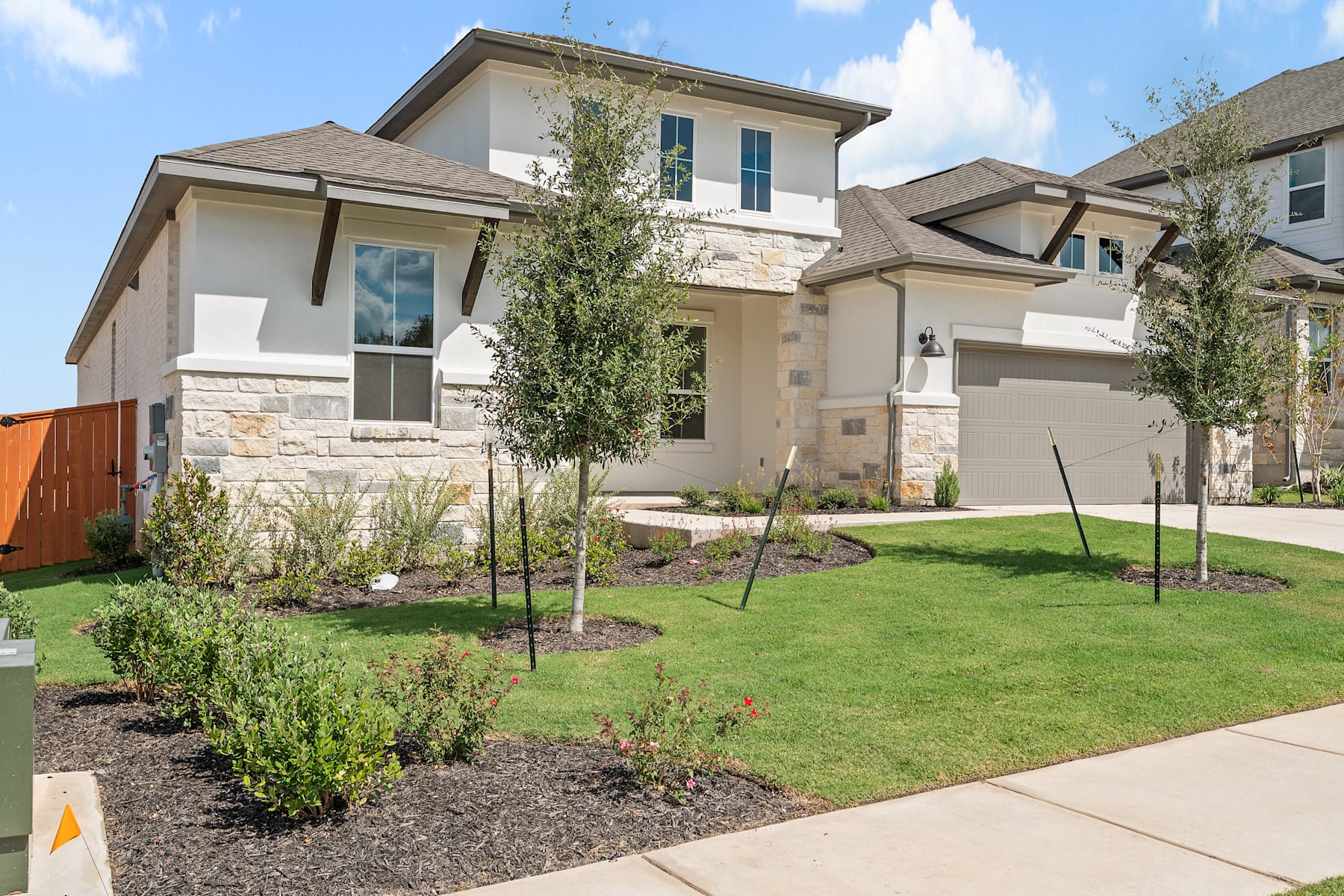 A well-manicured residential neighborhood with a two-story house featuring a stone exterior, a lush green lawn, and various landscaping elements such as trees and shrubs.