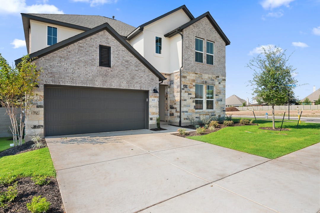 A two-story residential house with a gray exterior, a garage, and a well-manicured lawn in the foreground, set against a backdrop of blue sky and lush greenery.