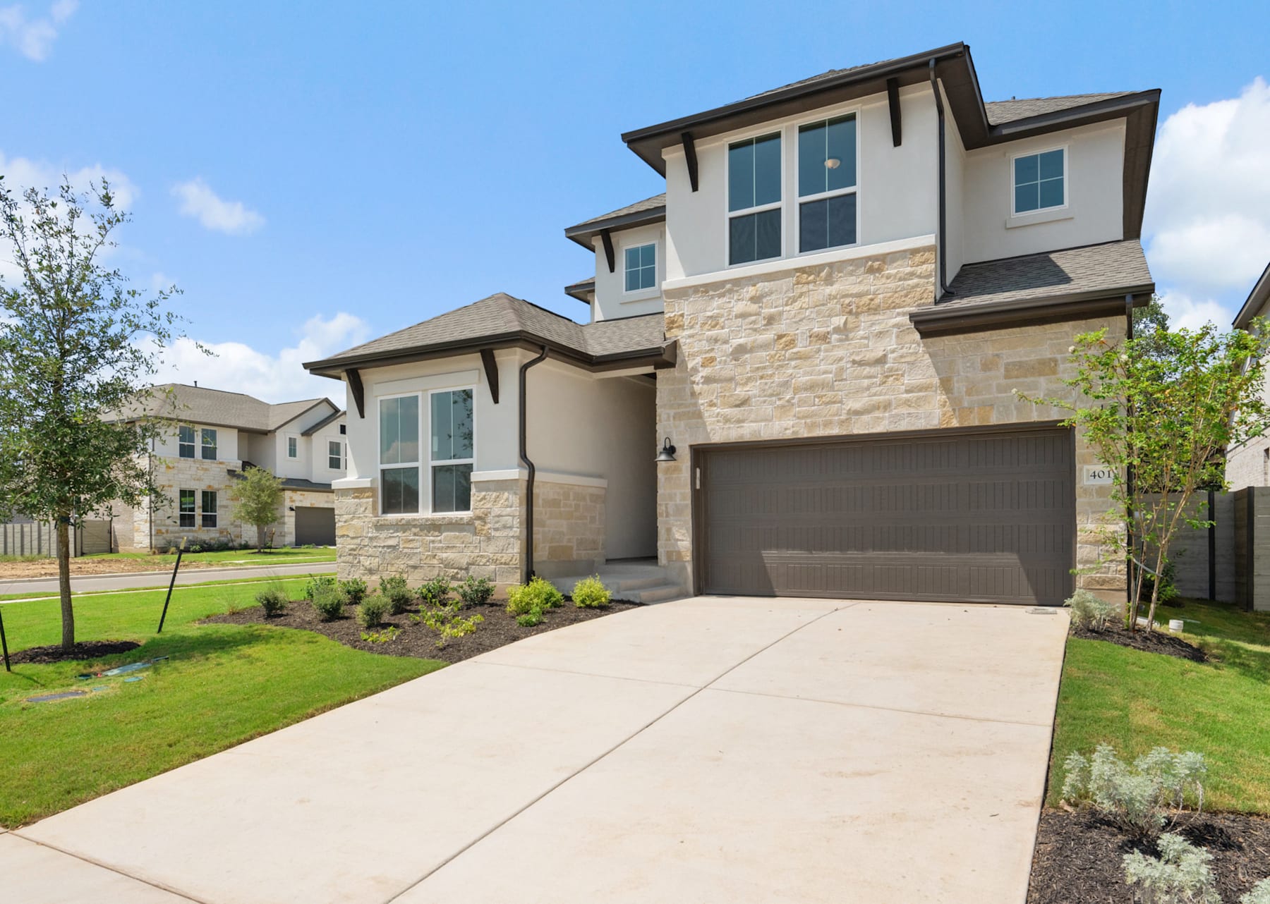 A two-story modern house with a stone exterior, a garage, and a paved driveway surrounded by lush greenery and a clear blue sky.