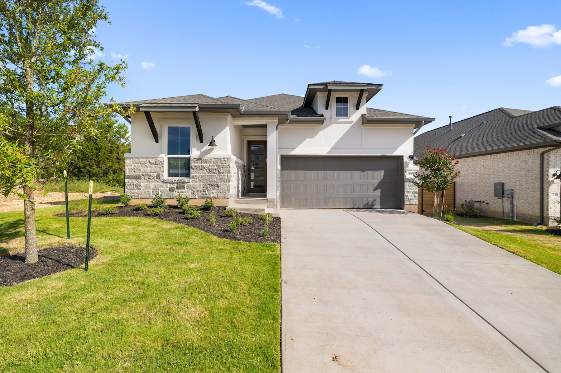 A modern two-story house with a stone exterior, surrounded by a well-manicured lawn and lush greenery, set against a clear blue sky with scattered clouds.