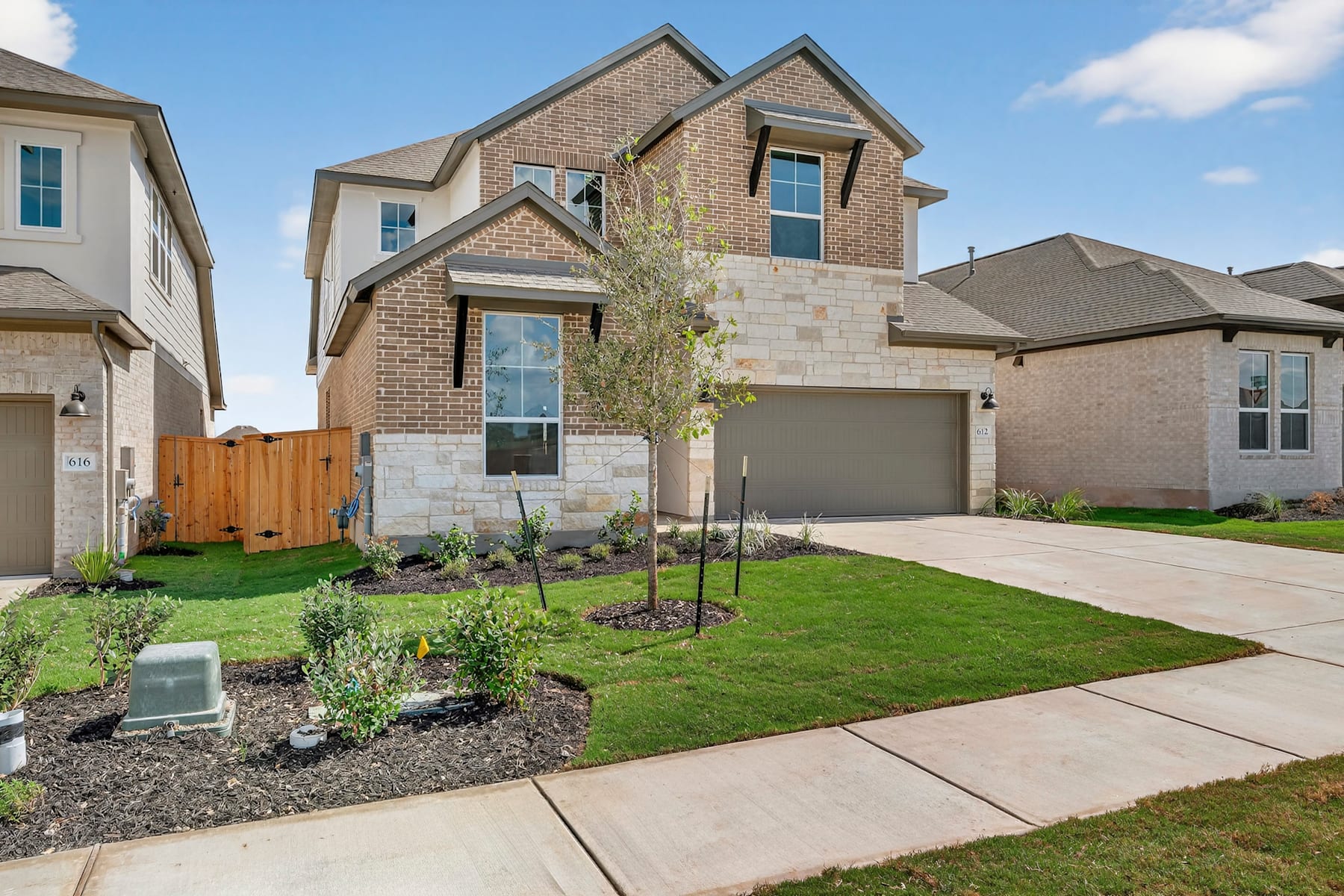 A two-story brick house with a well-manicured lawn, surrounded by other residential buildings, set against a clear blue sky with some clouds.