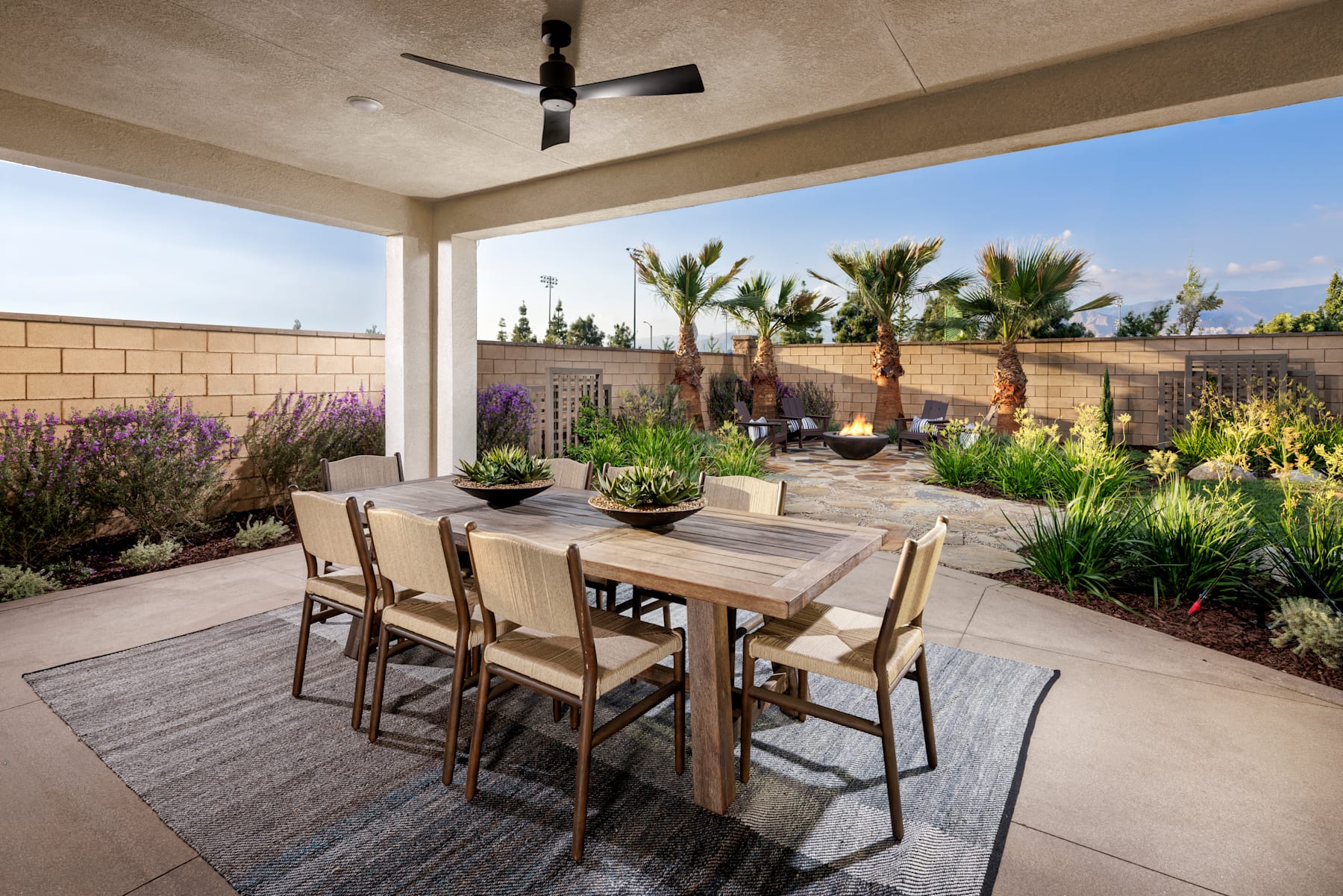 A covered outdoor dining area with a wooden table and chairs, surrounded by lush landscaping featuring palm trees, flowering plants, and a brick wall in the background.