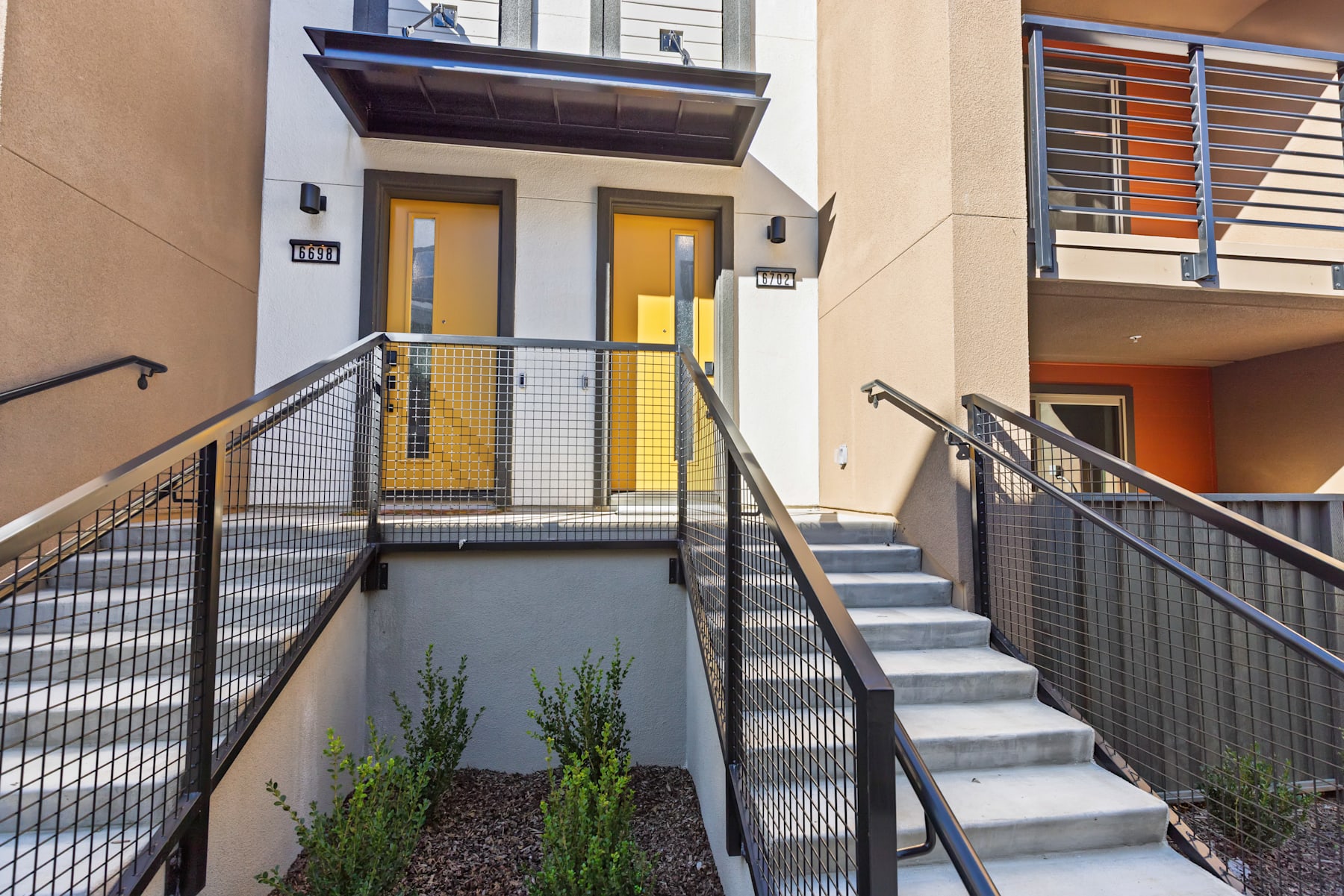 The image shows a modern, multi-story residential building with a prominent entrance featuring a yellow door and glass panels. The foreground includes a set of stairs leading up to the entrance, surrounded by a metal railing and landscaping elements.