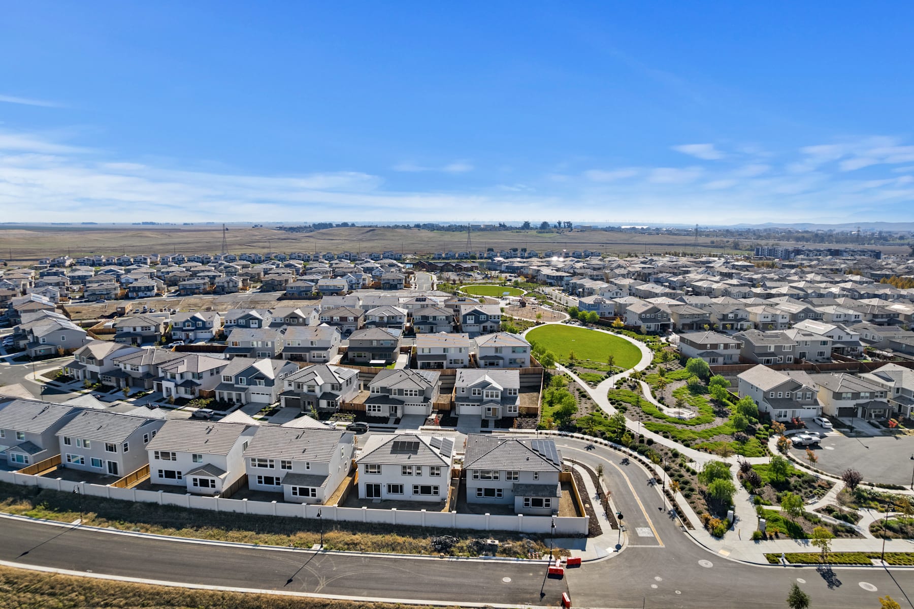 A sprawling residential neighborhood with rows of houses and a winding path surrounded by greenery, set against a backdrop of distant hills and a clear blue sky.