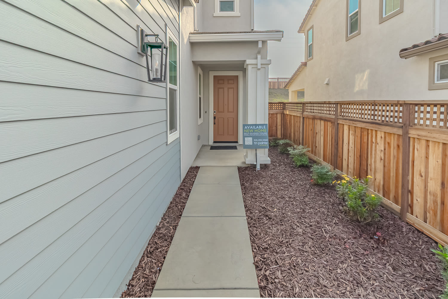 A narrow walkway lined with gravel leads to a wooden door, with a gray siding exterior and a lattice fence on the right side of the image.