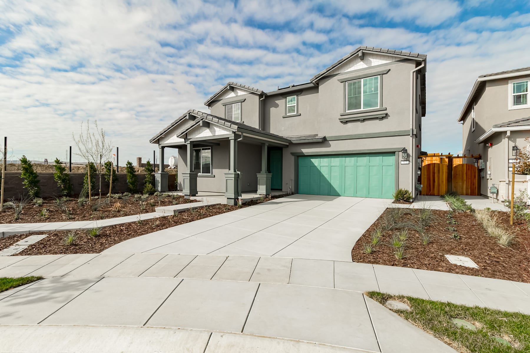 A modern two-story residential home with a gray exterior, a green garage door, and a paved walkway leading to the front entrance, set against a backdrop of a cloudy blue sky.