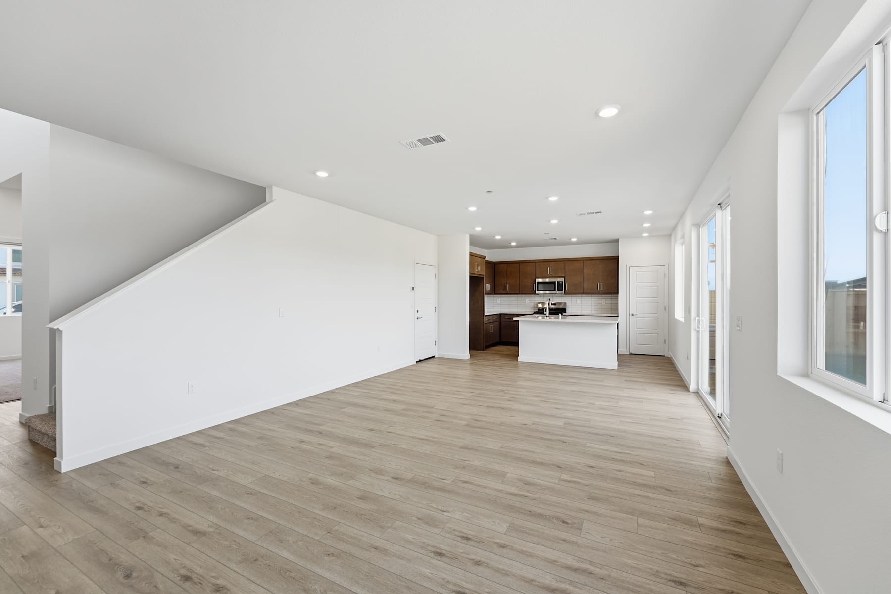 A spacious, open-concept living area with a modern kitchen visible in the background, featuring white walls, hardwood flooring, and recessed lighting.