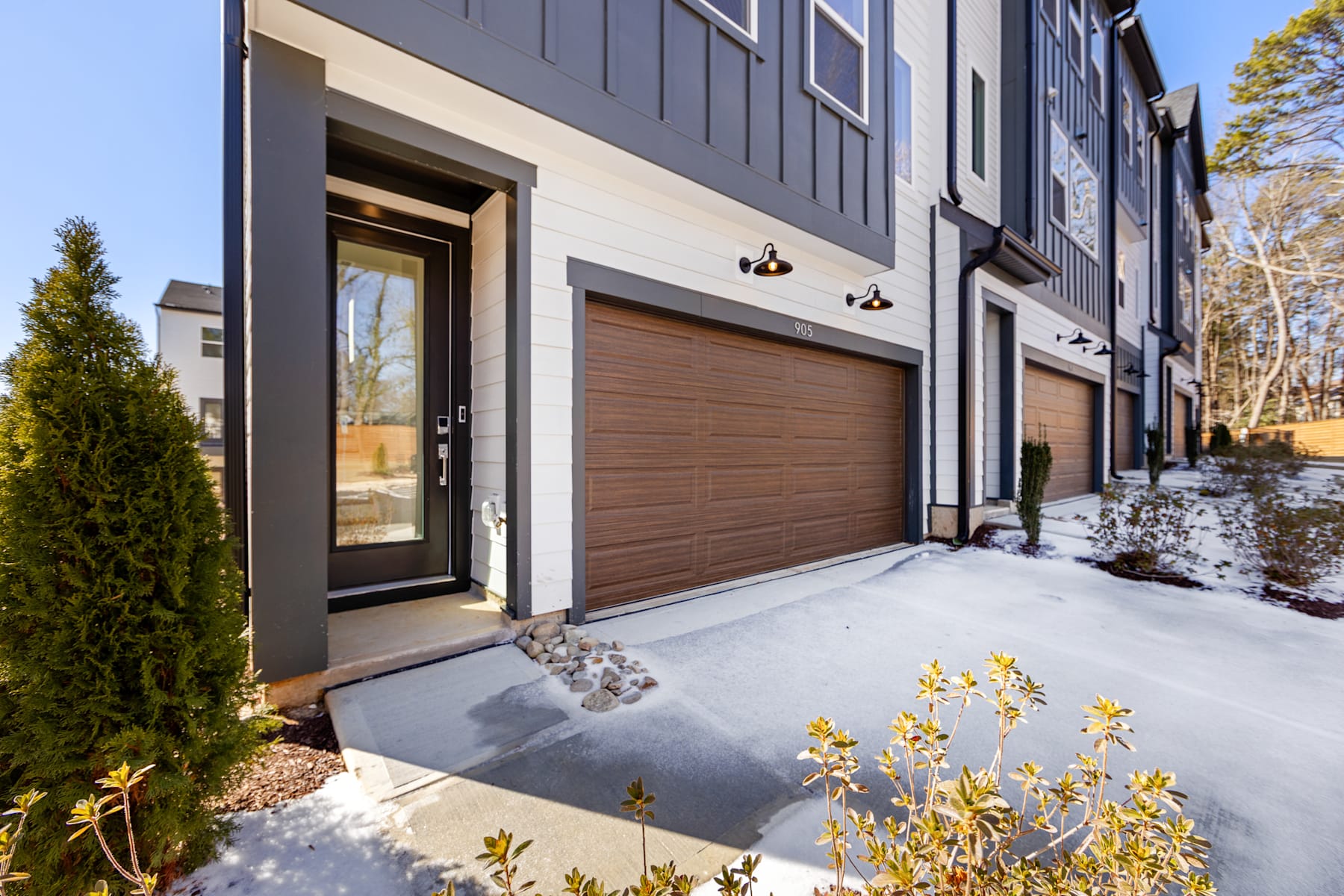 A modern townhouse with a wooden garage door, surrounded by snow-covered ground and evergreen shrubs, set against a backdrop of a gray building facade.