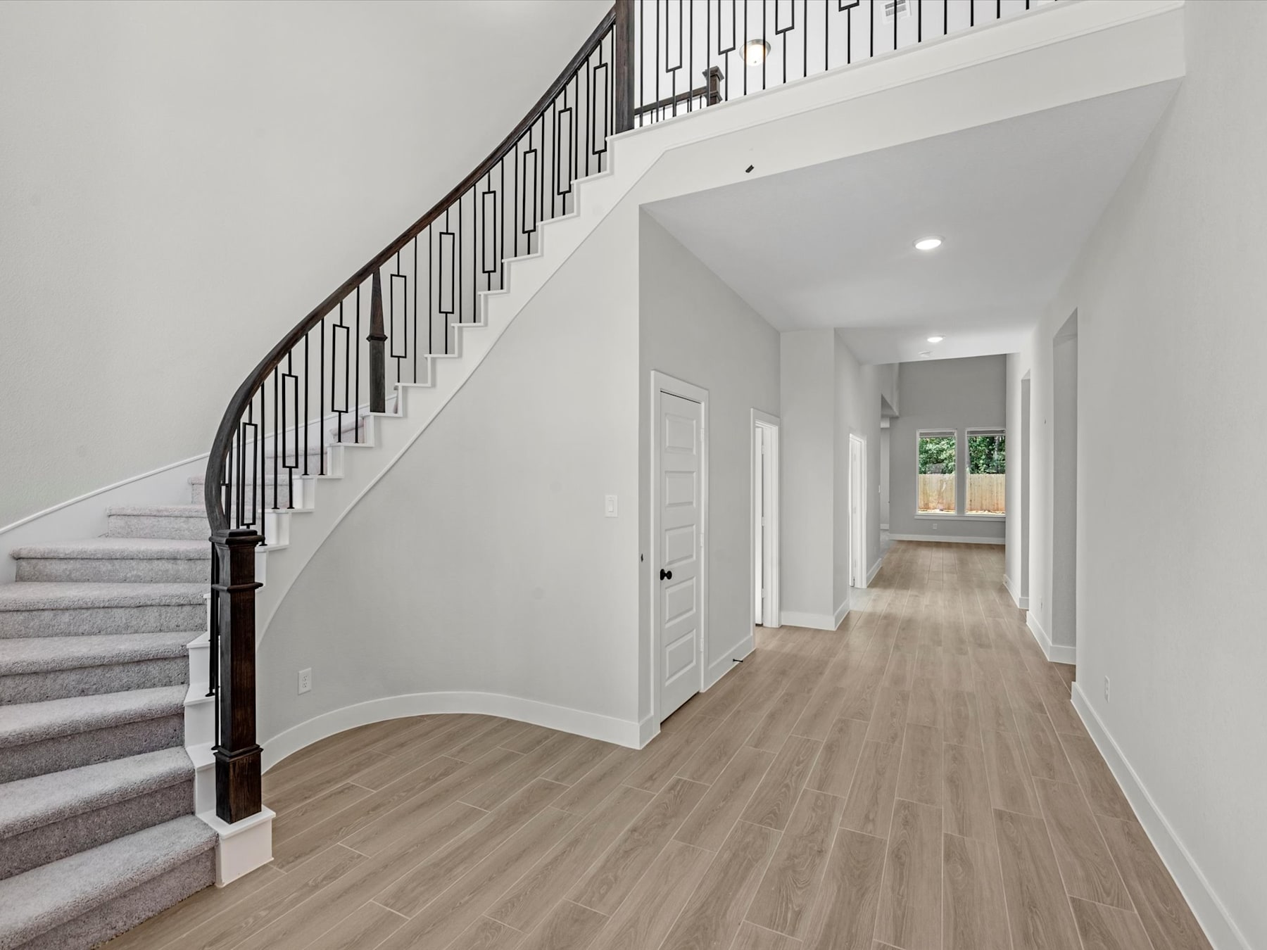 A spacious and well-lit hallway with a wooden staircase and hardwood flooring, leading to a doorway at the end of the corridor.
