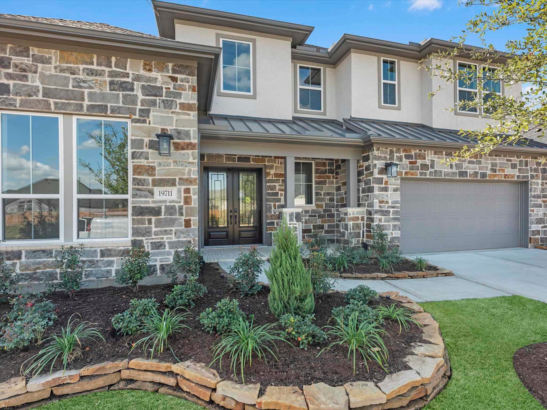 A two-story residential home with a stone exterior, a metal roof, and a well-landscaped front yard featuring a variety of plants and a stone border.