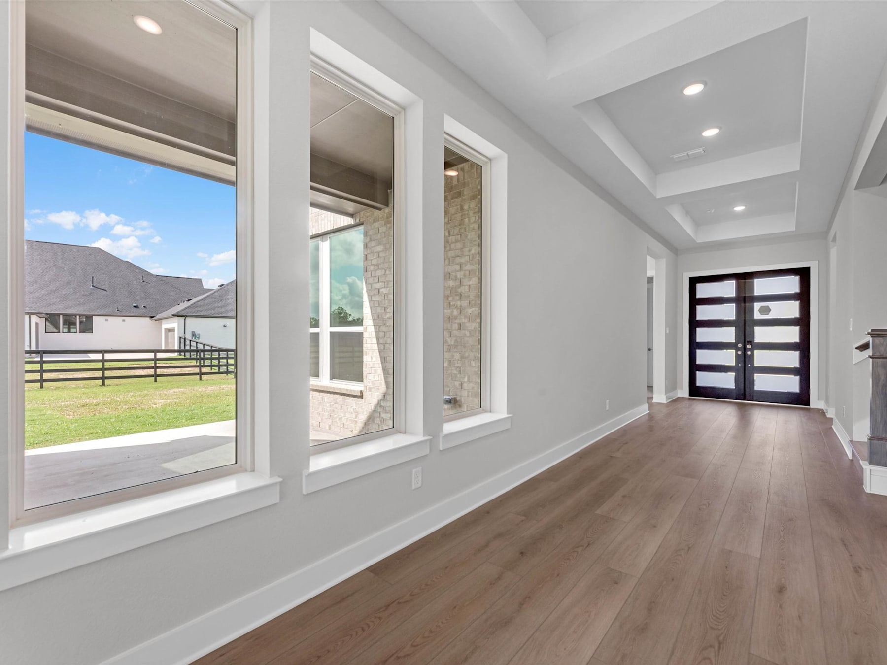 A spacious and bright interior with large windows overlooking a grassy outdoor area, featuring a wooden floor and a modern front door.