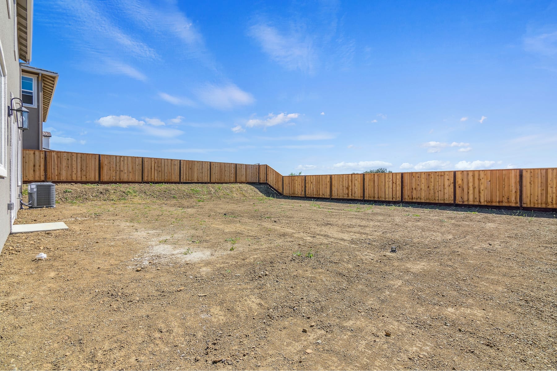 A wooden fence surrounds a barren, dirt-covered yard against a backdrop of a clear blue sky with scattered clouds.