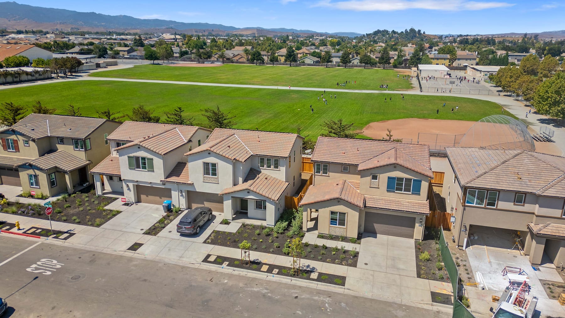 A residential neighborhood with rows of houses and a large grassy field in the background, surrounded by mountains and a blue sky.