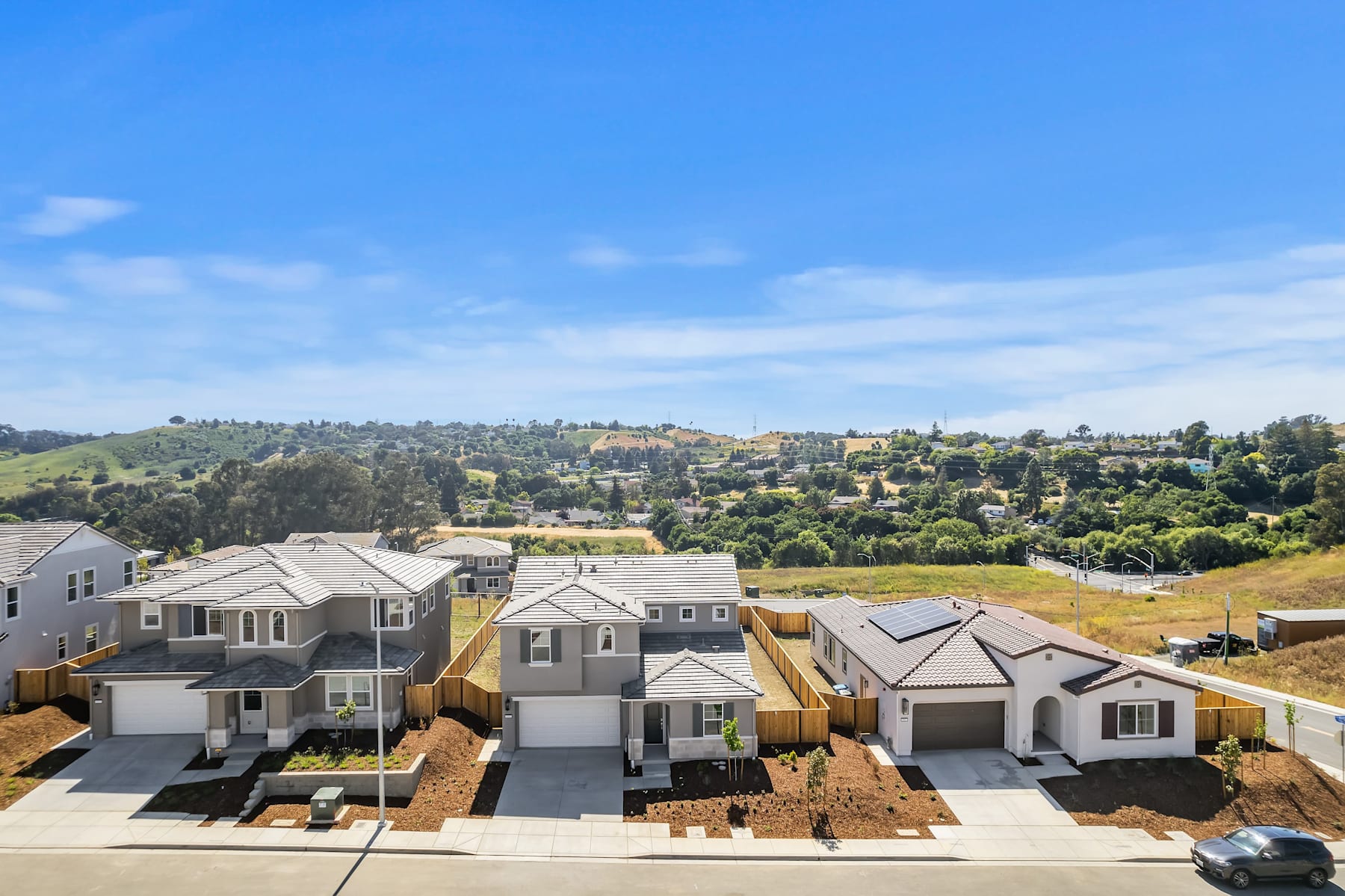 A residential neighborhood with rows of houses nestled among rolling hills, set against a clear blue sky with scattered clouds.