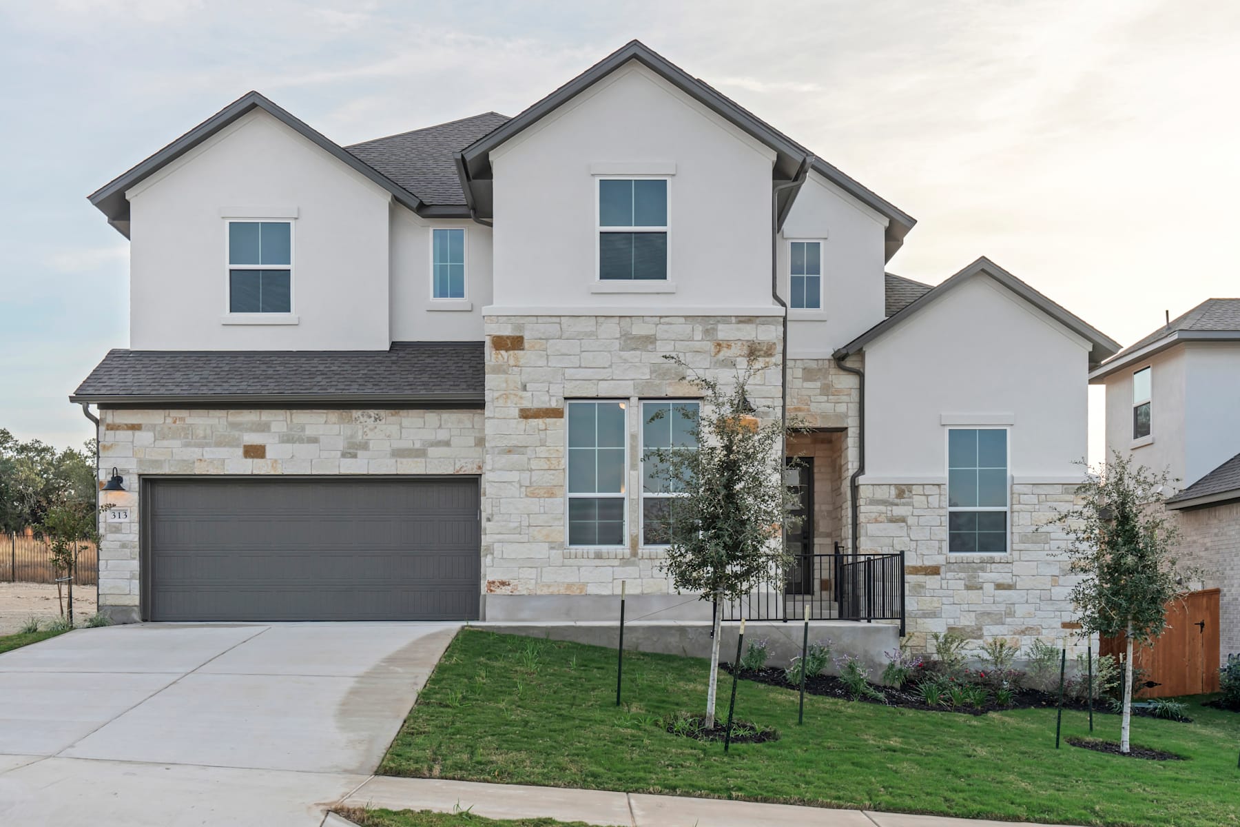 A two-story white and stone house with a garage, surrounded by a grassy lawn and landscaping, set against a cloudy sky.