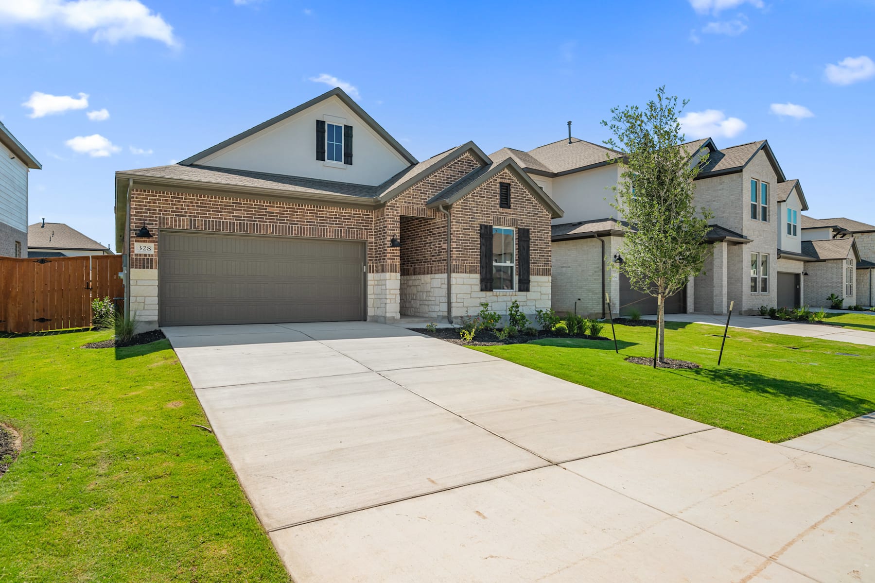 A newly constructed two-story brick house with a garage, surrounded by a well-manicured lawn and trees, set against a blue sky with scattered clouds.