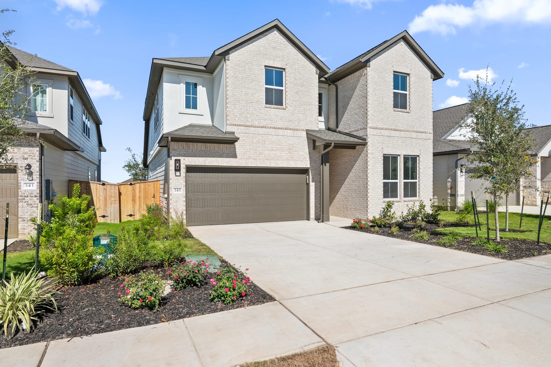 A modern two-story residential house with a well-landscaped front yard, featuring a concrete driveway and a variety of plants and flowers.