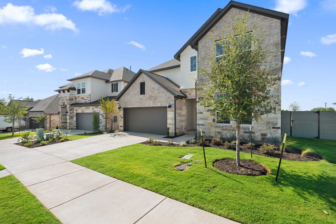 A well-manicured lawn and landscaping surround a large, multi-story residential building with a stone and siding exterior, featuring a garage and multiple windows against a backdrop of a blue sky with scattered clouds.