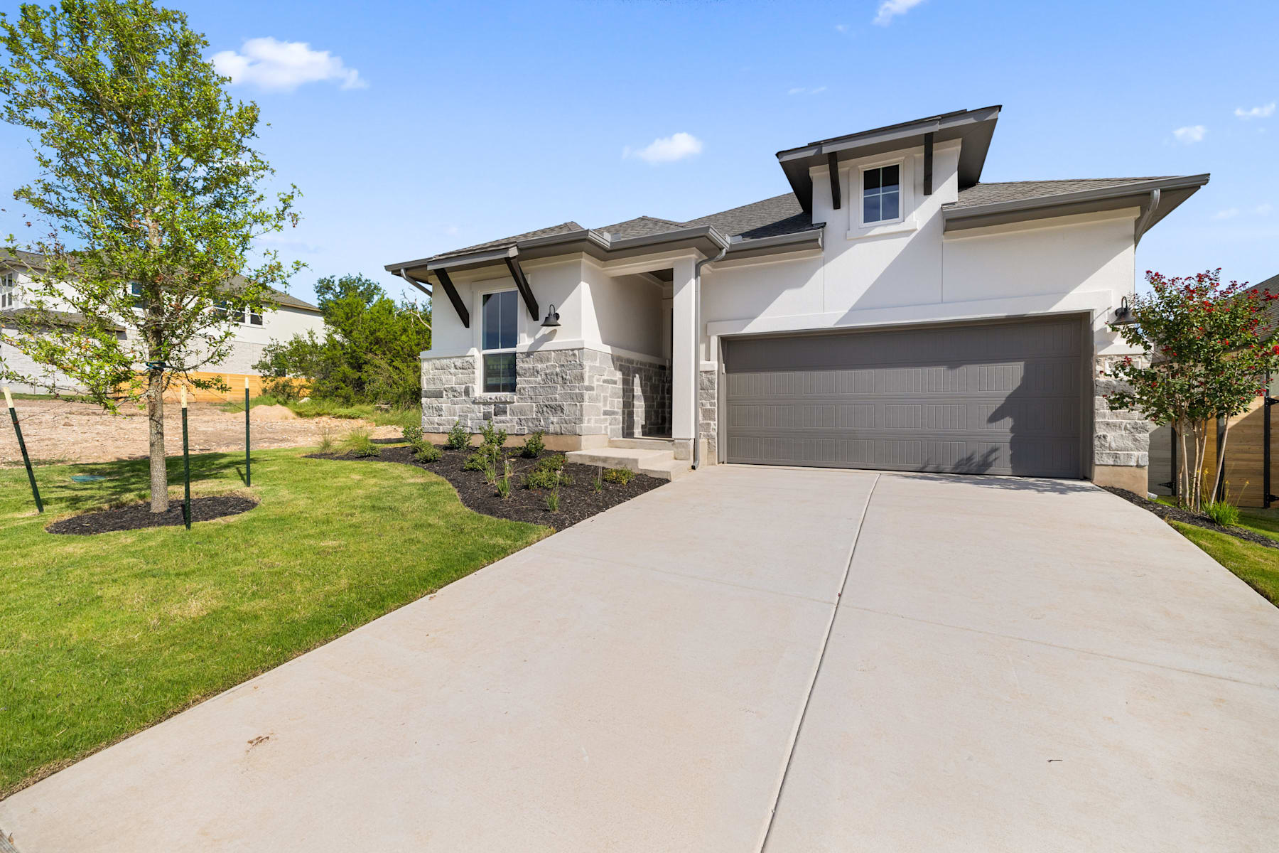 A modern two-story house with a stone exterior, a garage, and a well-maintained lawn in the foreground, set against a backdrop of lush greenery and a clear blue sky.