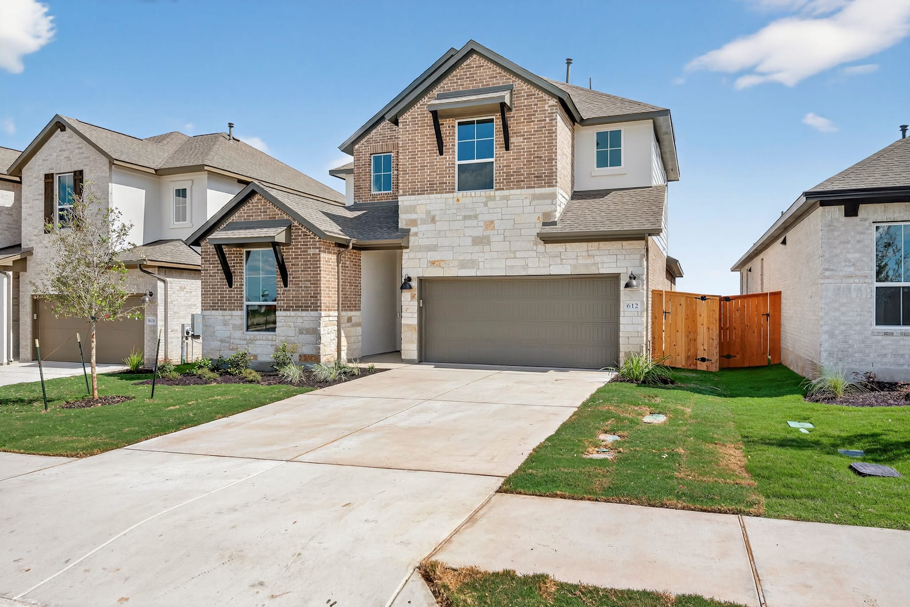 A two-story brick and stone house with a garage, surrounded by a well-manicured lawn and other similar houses in the background, set against a blue sky with scattered clouds.