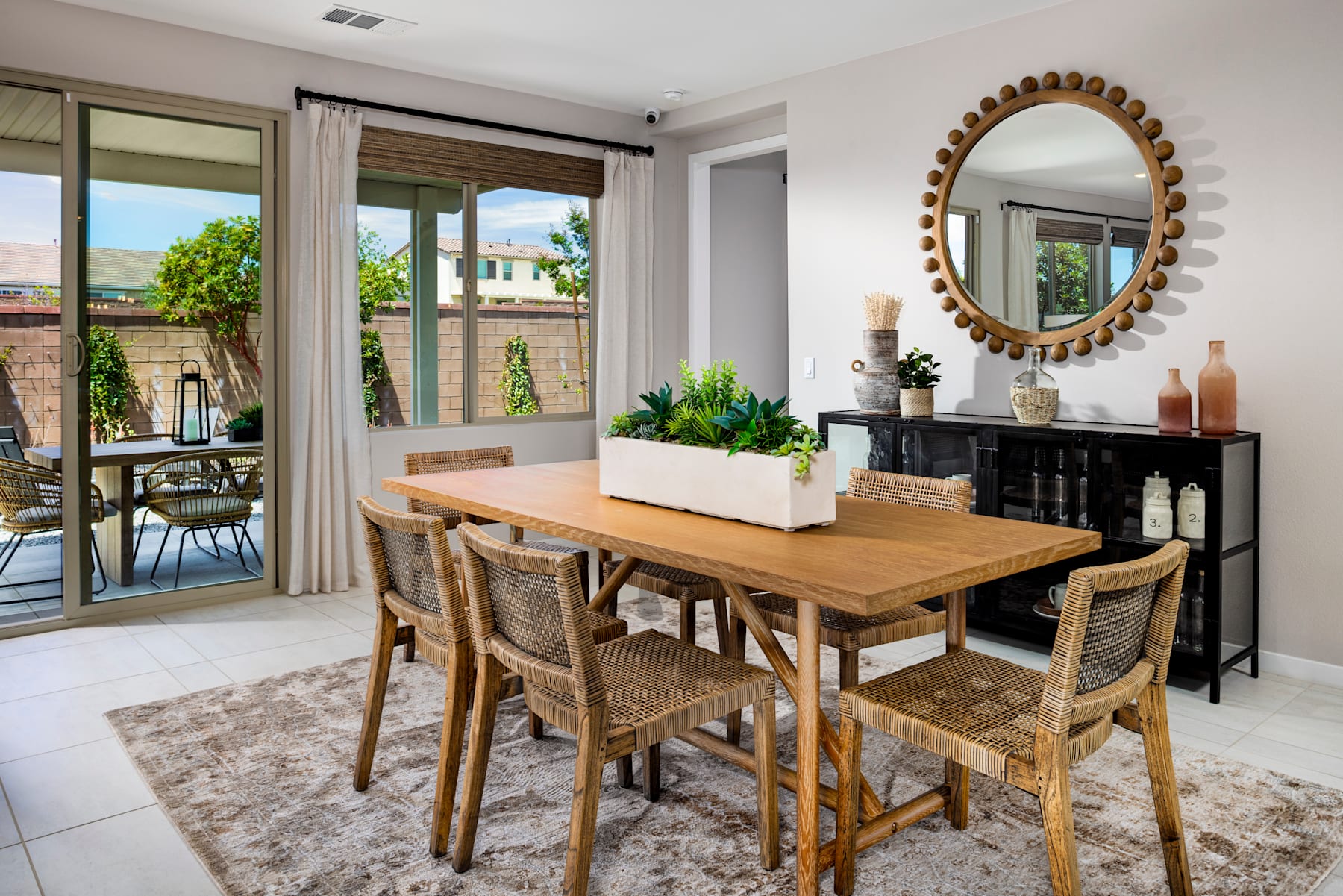 A cozy dining area with a wooden table and wicker chairs, surrounded by large windows overlooking a lush outdoor space, and accented by a decorative mirror and various potted plants.