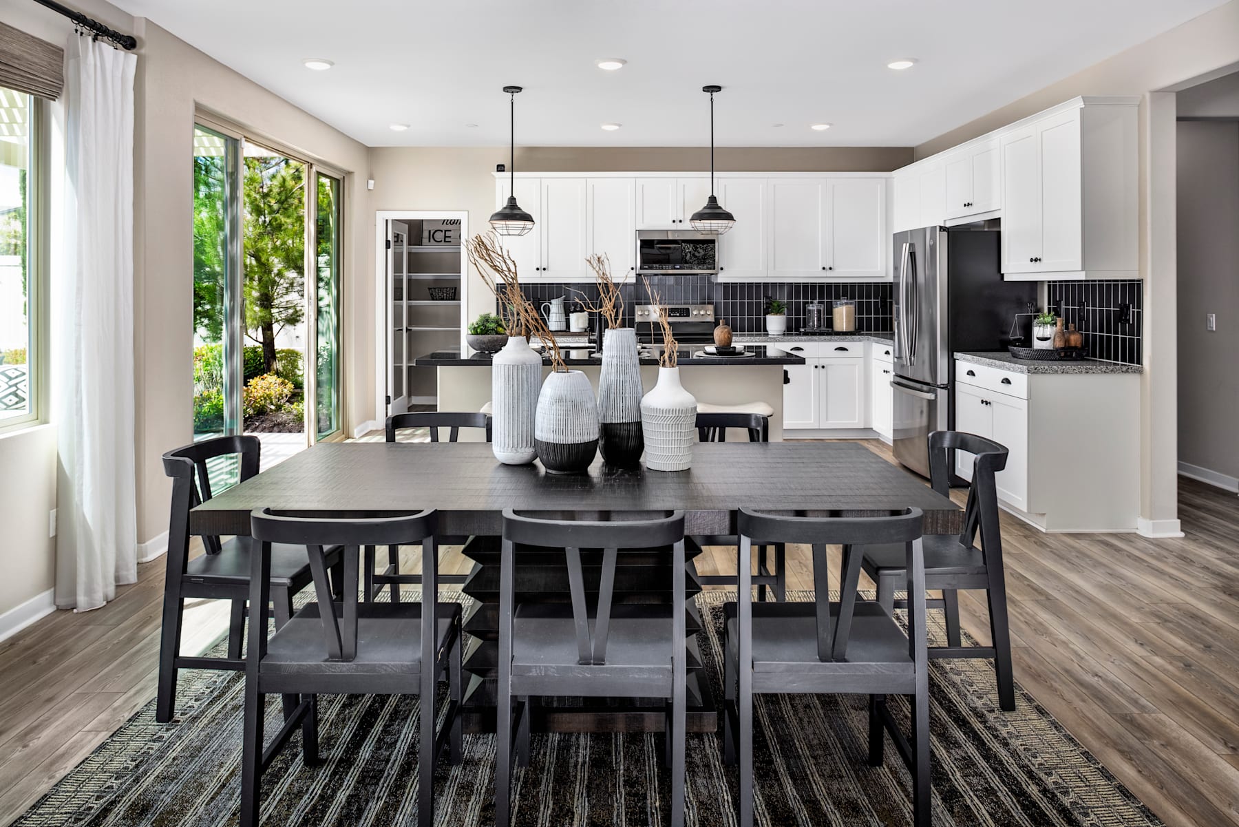 A modern and spacious kitchen with a large dining table, dark wooden chairs, and a view of the lush greenery outside through the large windows.