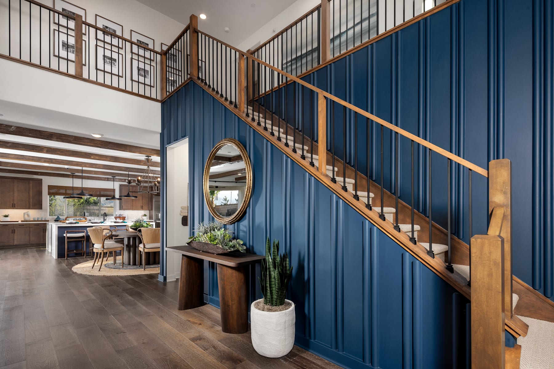 A modern and stylish interior with a wooden staircase, a round mirror, and potted plants decorating the entryway, leading to an open-concept living space with a kitchen visible in the background.