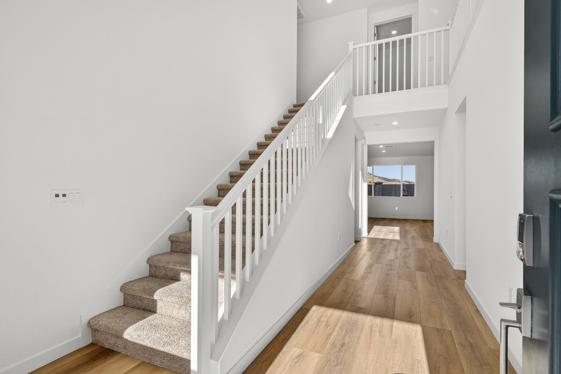 A bright and spacious hallway with a wooden staircase and white railings leading up to the second floor, with a hardwood floor extending into the background.