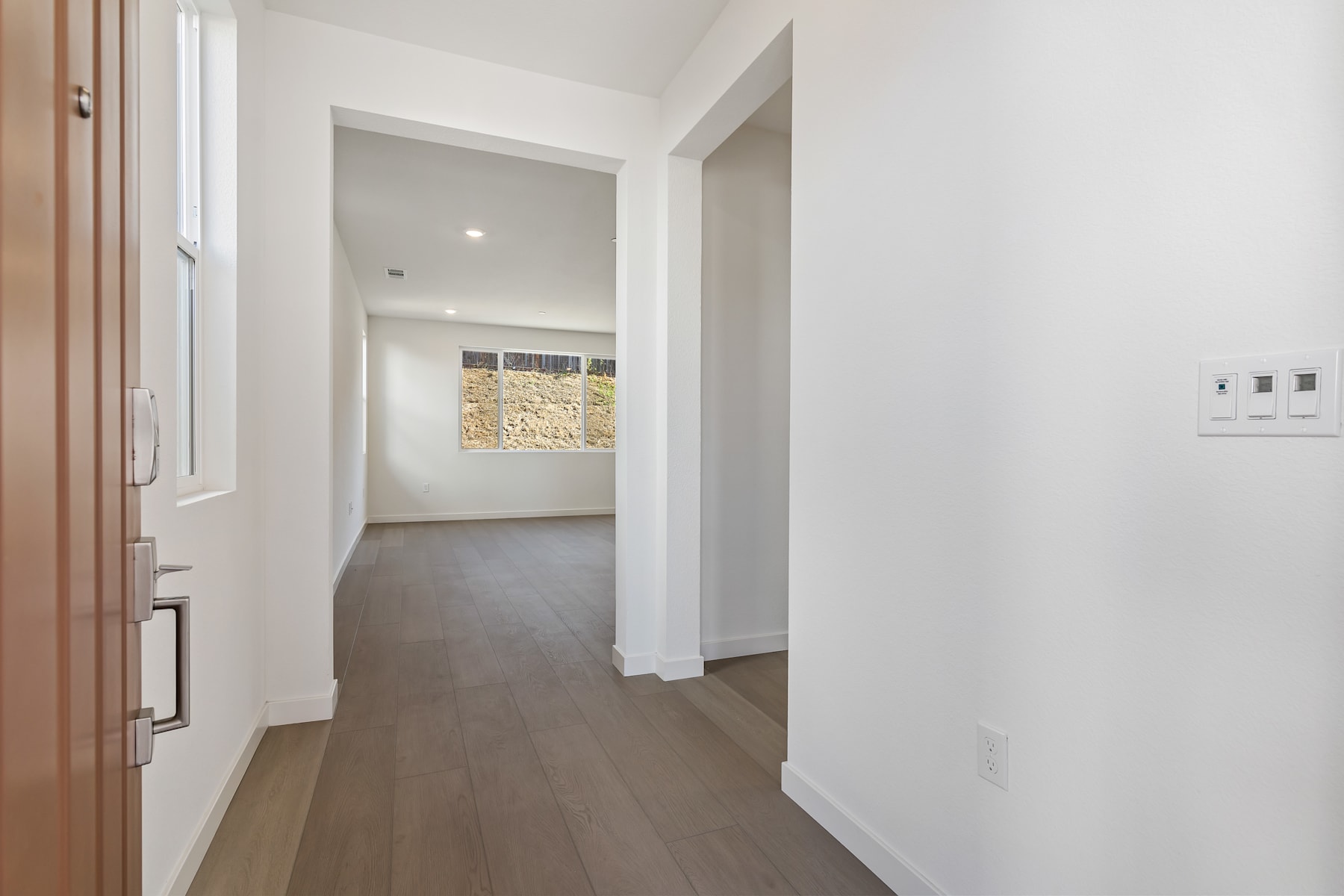 A bright, minimalist hallway with wooden floors and white walls, leading to a room with a window.
