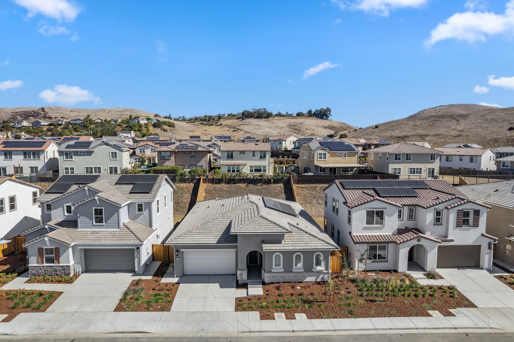 A residential neighborhood with rows of modern, two-story houses nestled among rolling hills and a clear blue sky with scattered clouds.