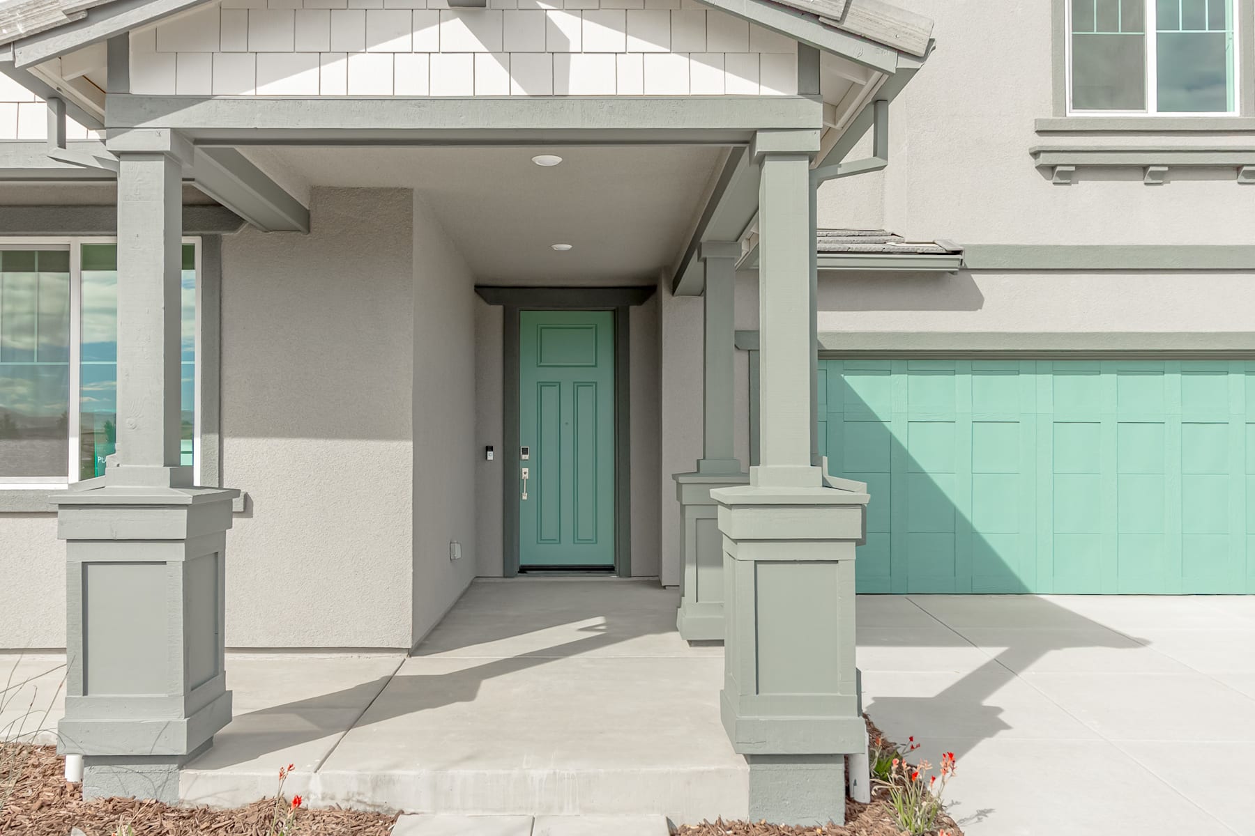 A modern, two-story house with a covered porch, gray siding, and a teal-colored front door, surrounded by a well-maintained landscaped area.