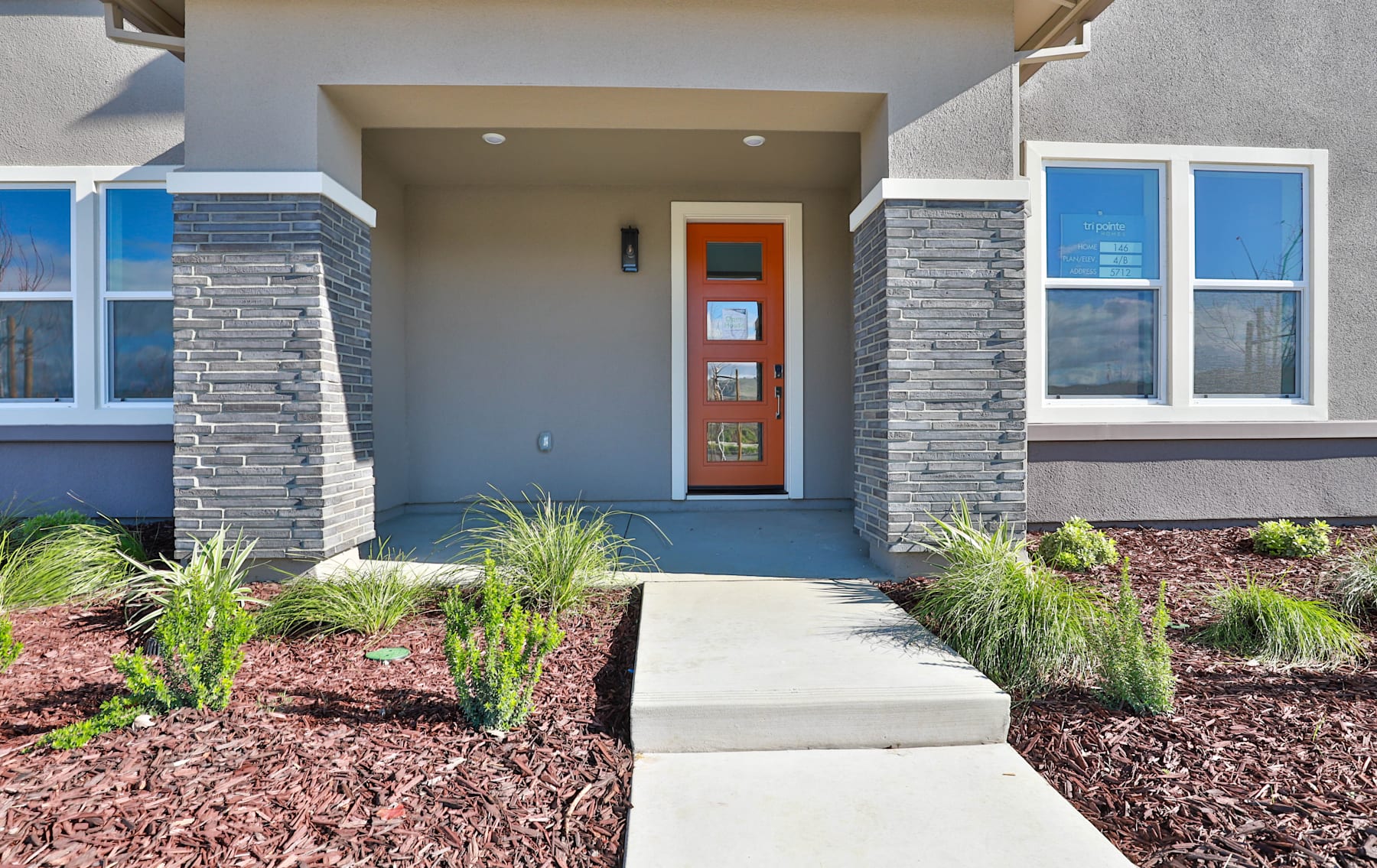 The image shows the front entrance of a modern, two-story residential home with a stone facade, a red front door, and a paved walkway leading up to the entrance. The foreground features a landscaped area with plants and mulch, while the background includes a clear blue sky.