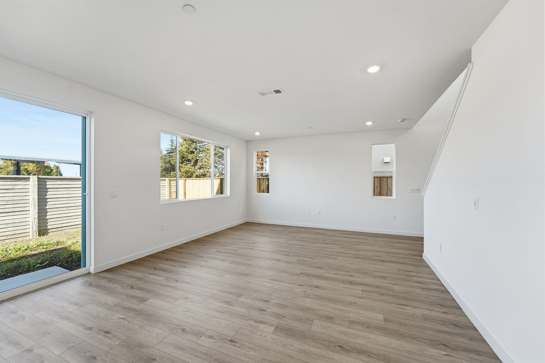 A spacious, well-lit room with a wooden floor, white walls, and large windows overlooking a backyard.