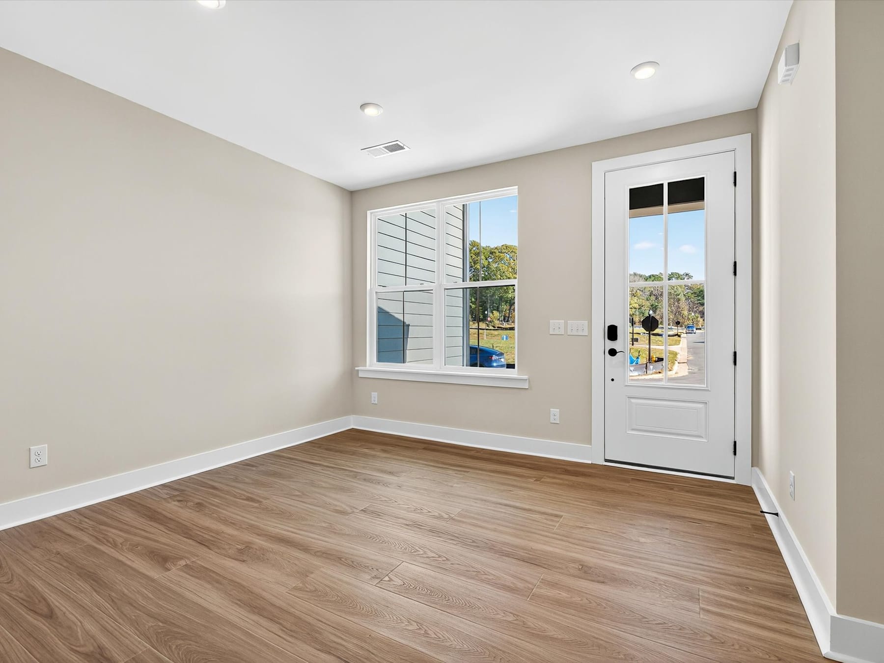A bright and spacious room with hardwood floors, a large window, and a white door leading to the exterior.