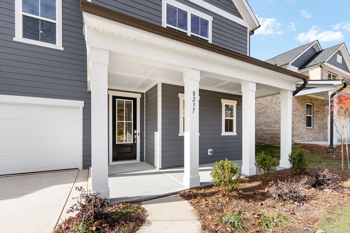 A modern two-story house with a covered porch, gray siding, and a white front door surrounded by landscaping in the foreground.