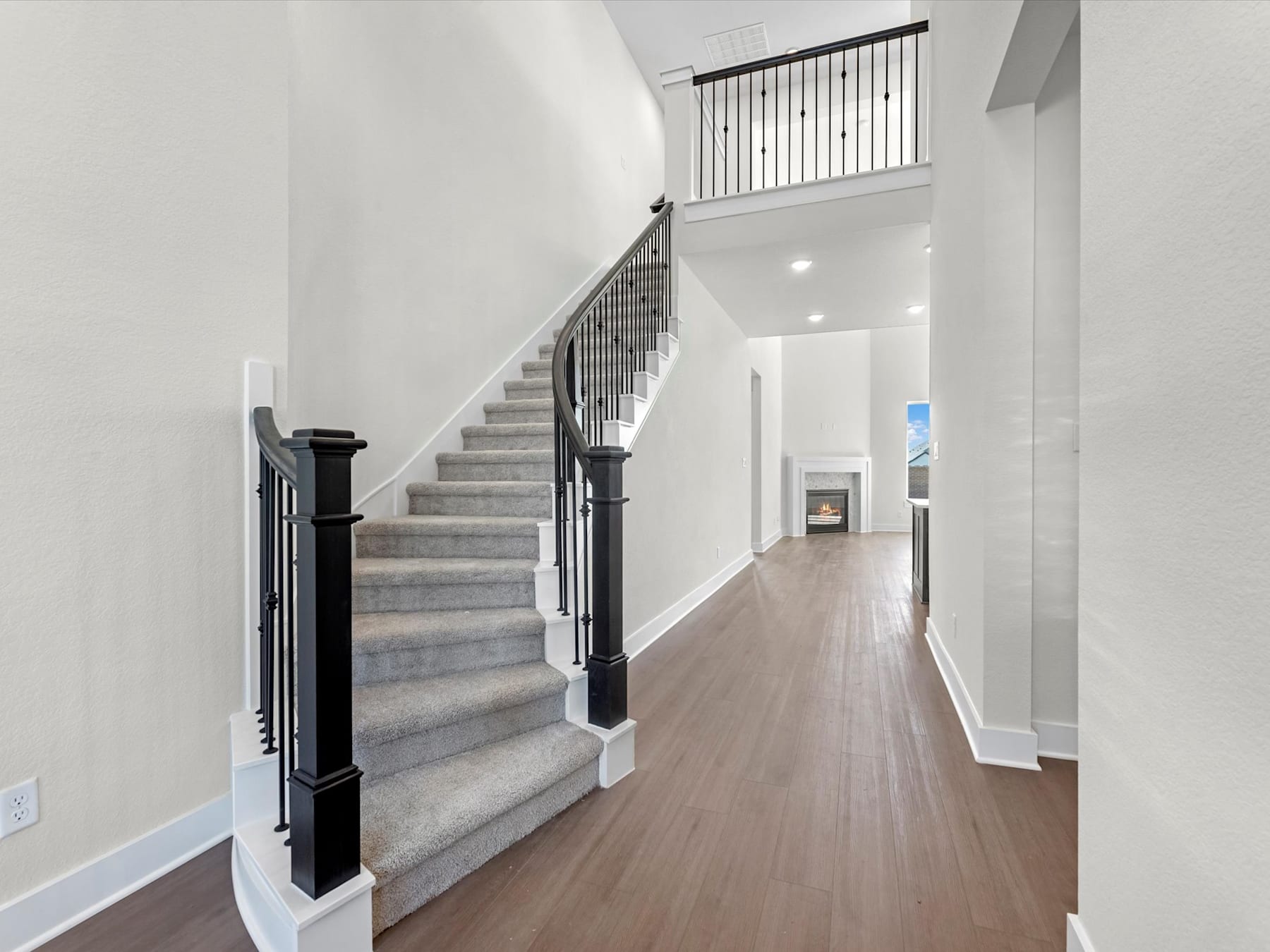 A spacious, well-lit hallway with a wooden floor, a staircase with a metal railing, and a doorway leading to another room.