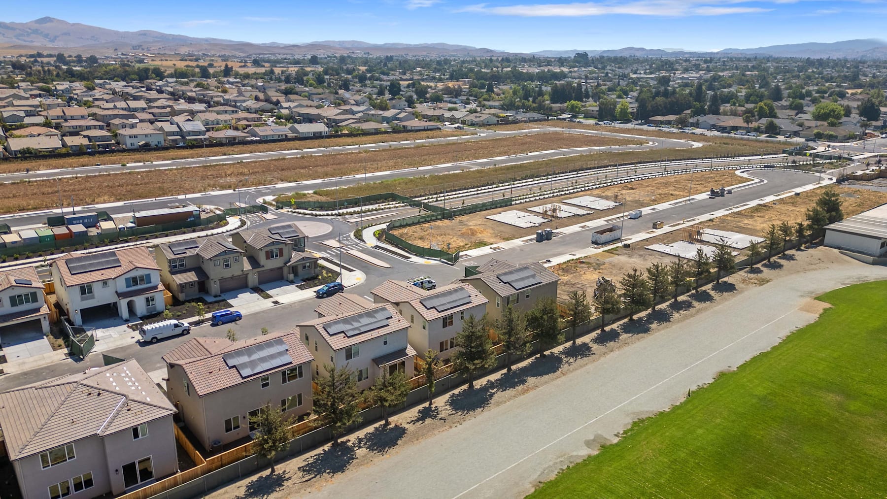 A residential neighborhood with rows of houses and apartment buildings, surrounded by a vast open field and mountains in the distance, under a clear blue sky.